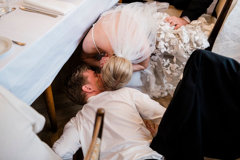 Bride and groom lying on floor under wedding table in playful candid moment during reception celebration.
