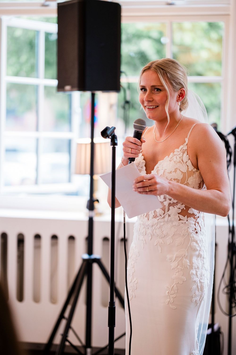 Smiling bride in lace wedding dress giving a speech at her wedding reception while holding a microphone and notes.