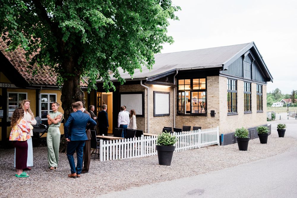 Wedding guests gathering outside a charming Danish venue with a large tree and white picket fence.