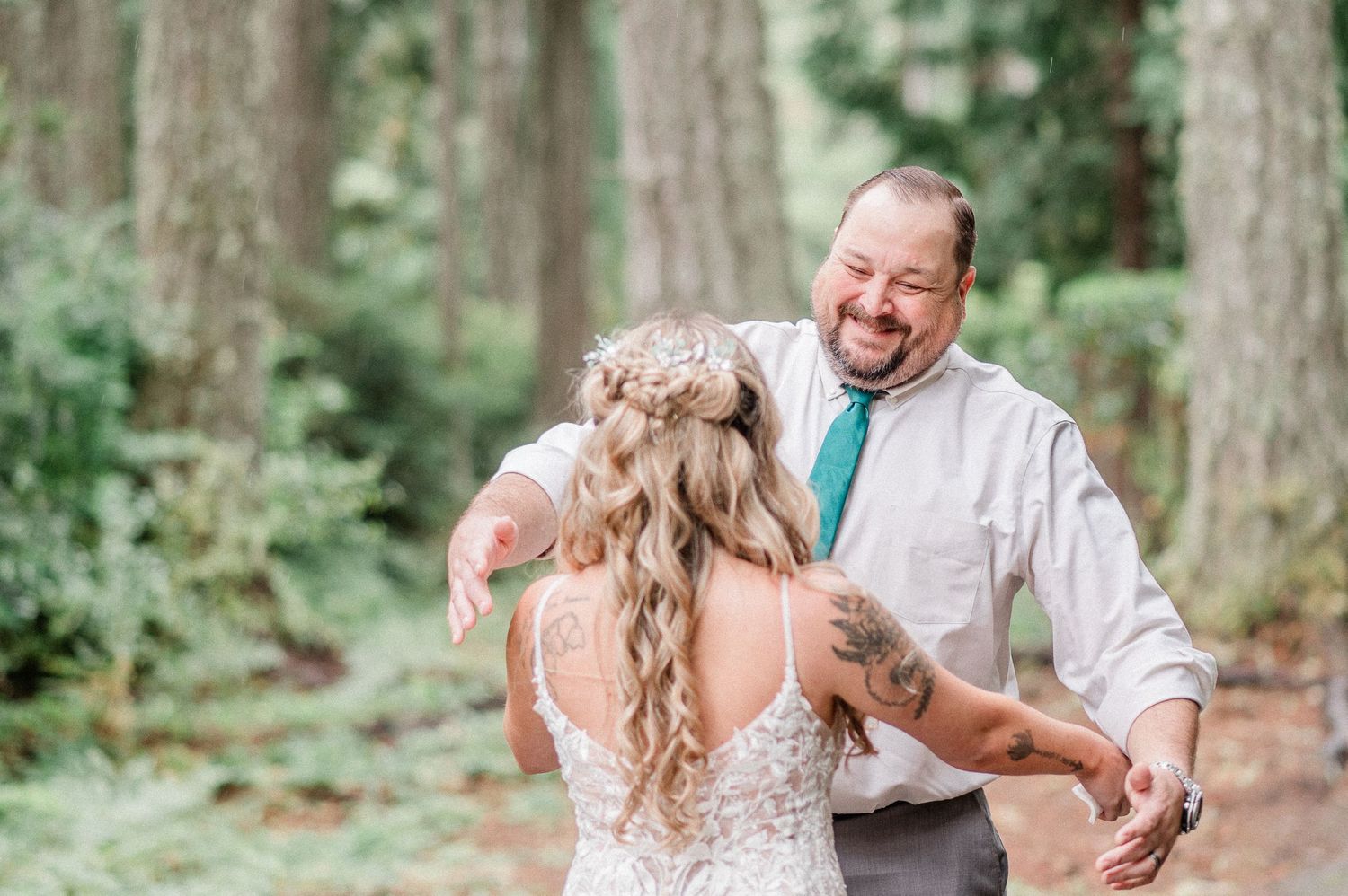 Bride and her father share a joyful first look moment in a lush forest setting, father smiling broadly in teal tie.