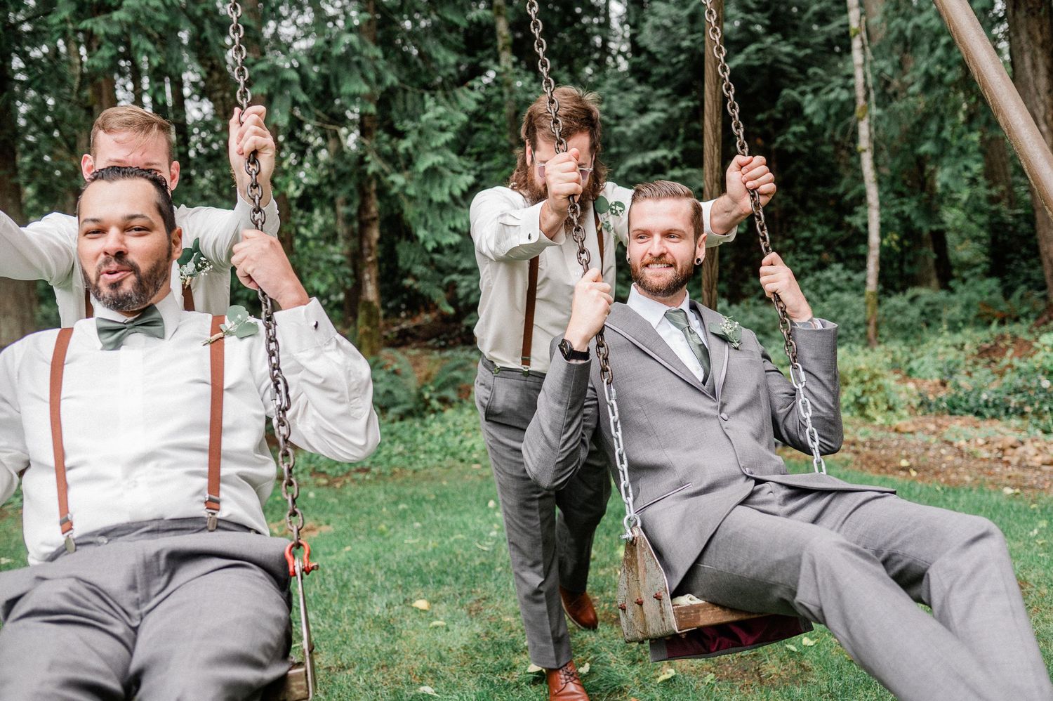 Groomsmen in gray suits and suspenders laughing and sitting on wooden swings outdoors at a wedding.