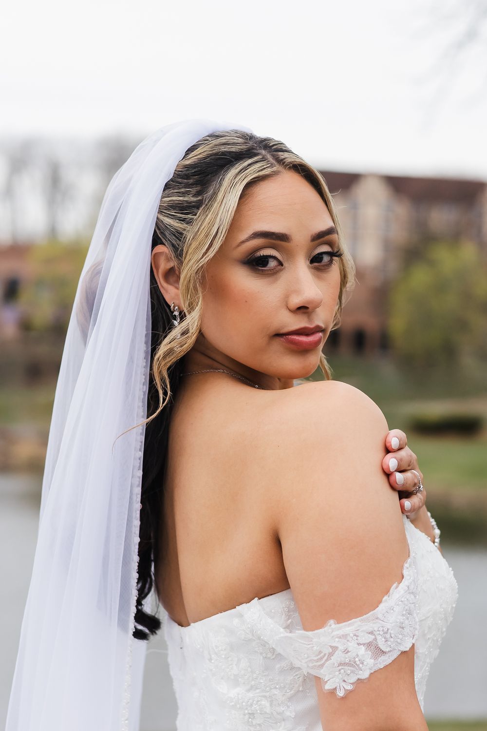 Bride in off-shoulder lace gown and white veil poses outdoors near water, looking over her shoulder.