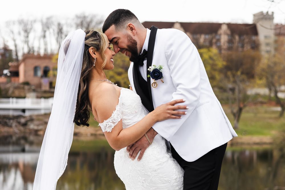 Bride and groom in white wedding attire share a romantic moment outdoors near a lake with autumn trees in background.