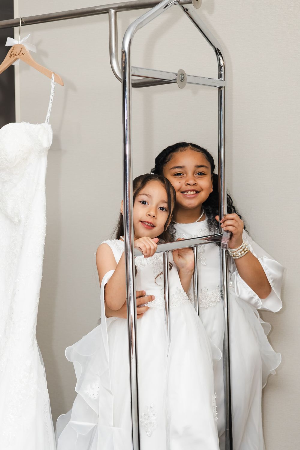 Two young girls in white dresses smile while posing near a clothing rack with a wedding gown in a bridal boutique.