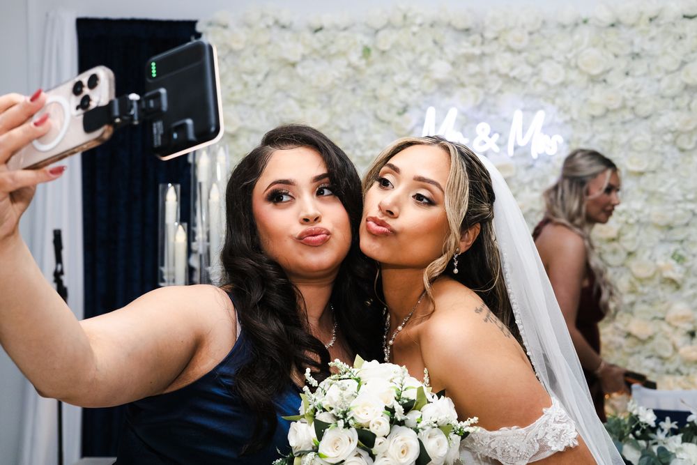 Bride in veil holding bouquet takes selfie with bridesmaid, both posing with pouts at wedding reception.