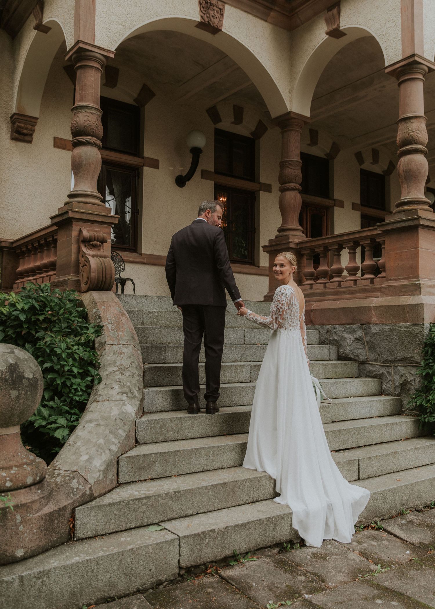 A bride in a white gown and groom in a dark suit hold hands ascending stone steps of an elegant building.
