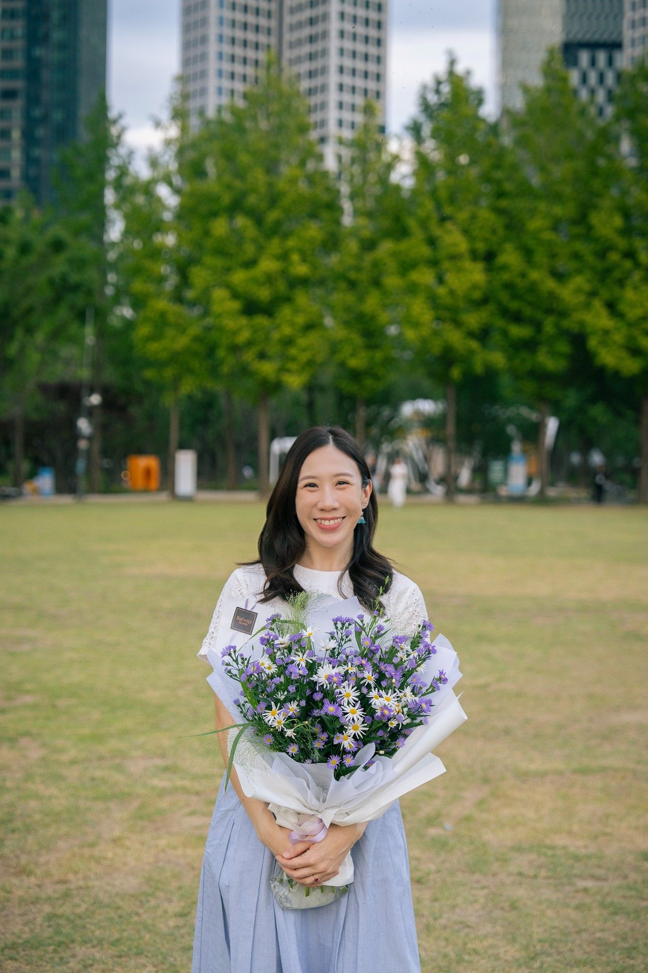 Smiling Asian woman holding a large purple and white flower bouquet in a green park with city skyscrapers behind her.
