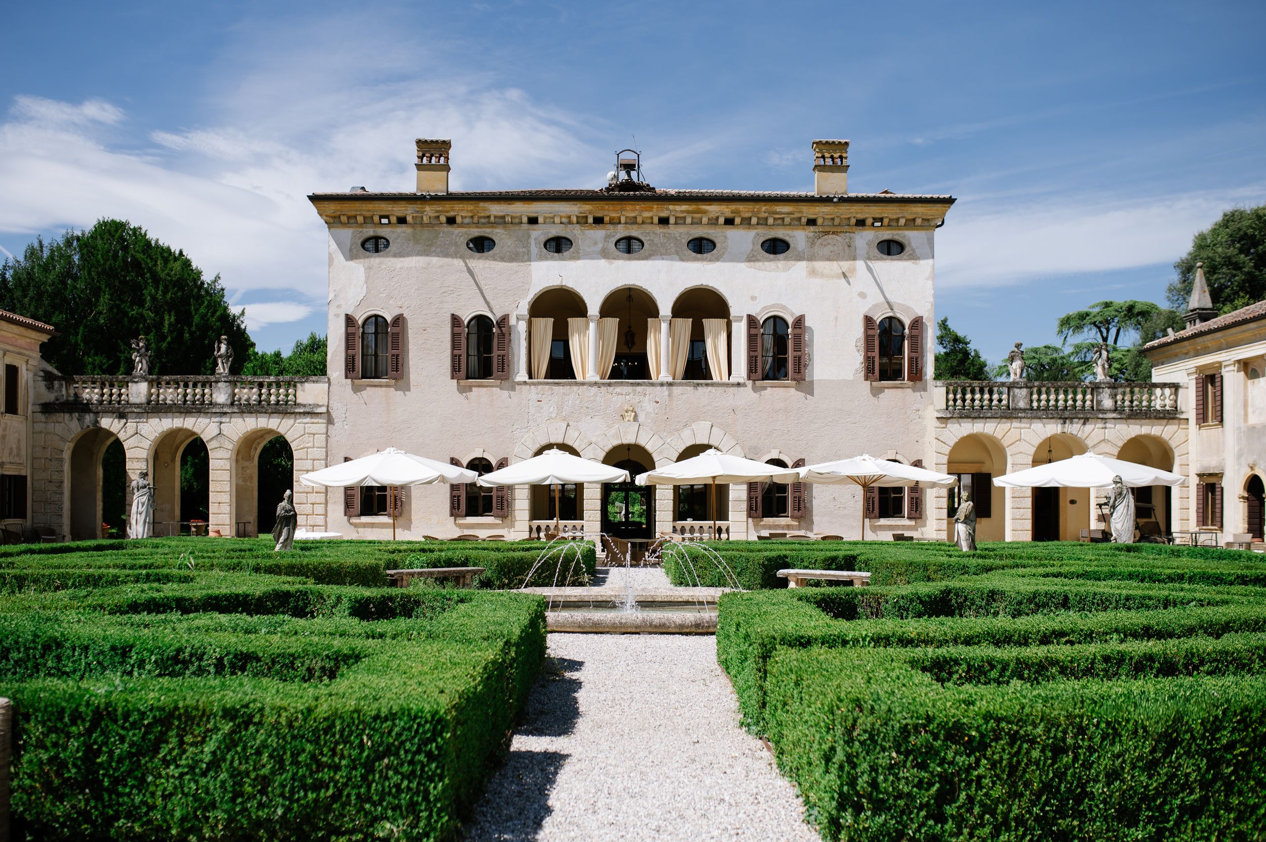 Elegant Italian villa with manicured box hedges, fountain, and arched loggia under blue sky.