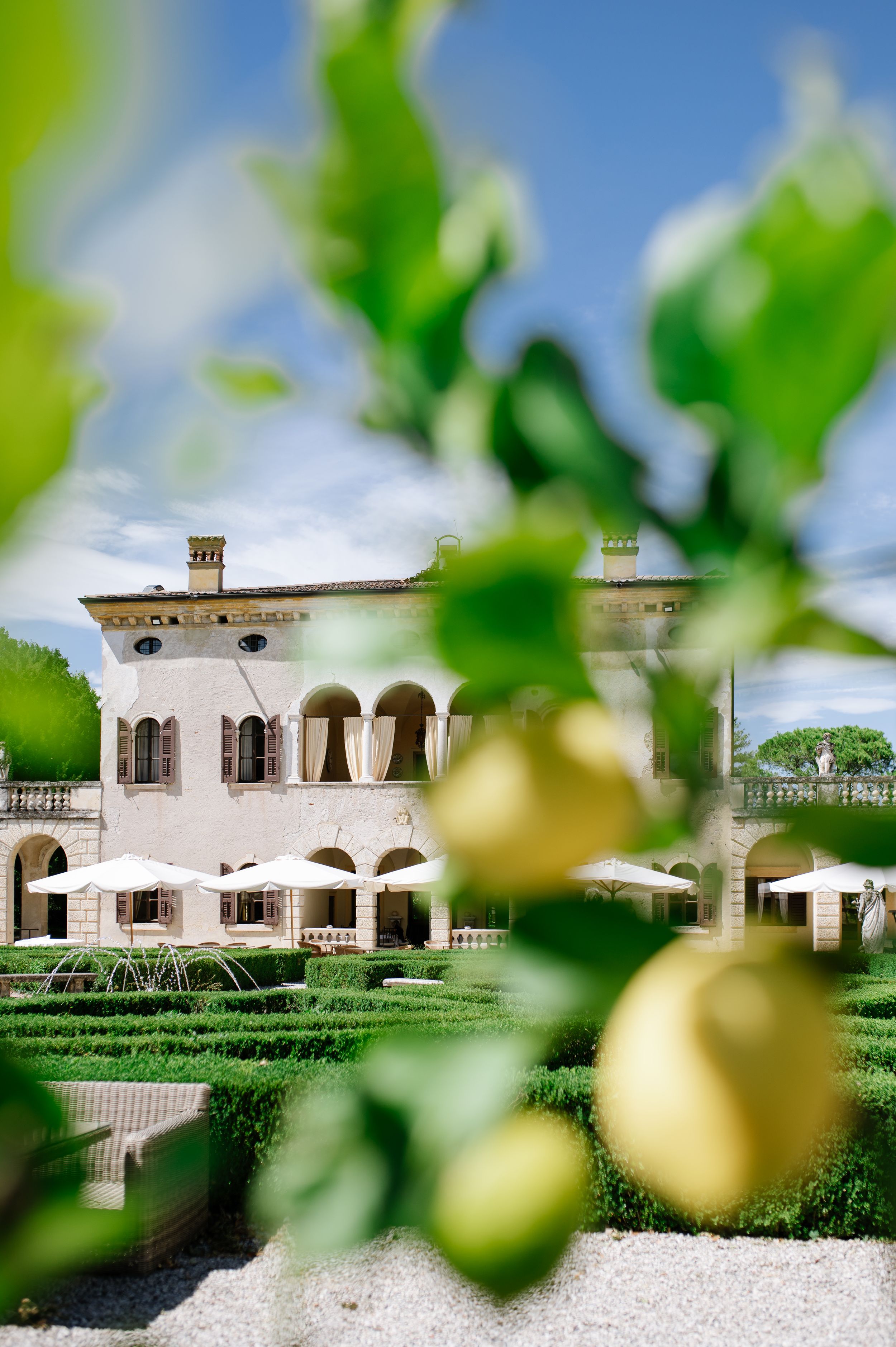 Elegant Italian villa wedding venue with manicured hedges, fountain, and lemon tree in foreground under blue sky.