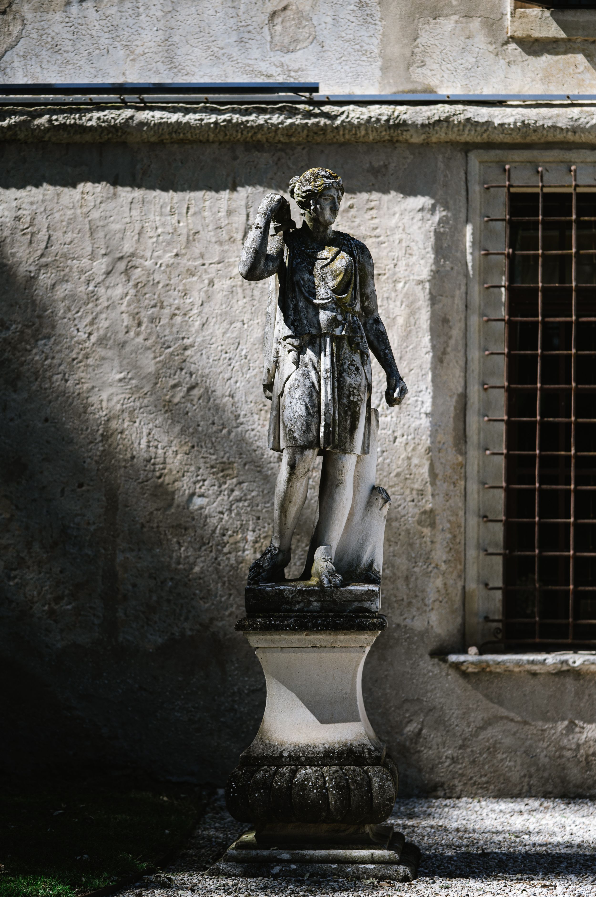 Ancient stone statue of a Roman figure on a pedestal against a weathered wall with barred window.