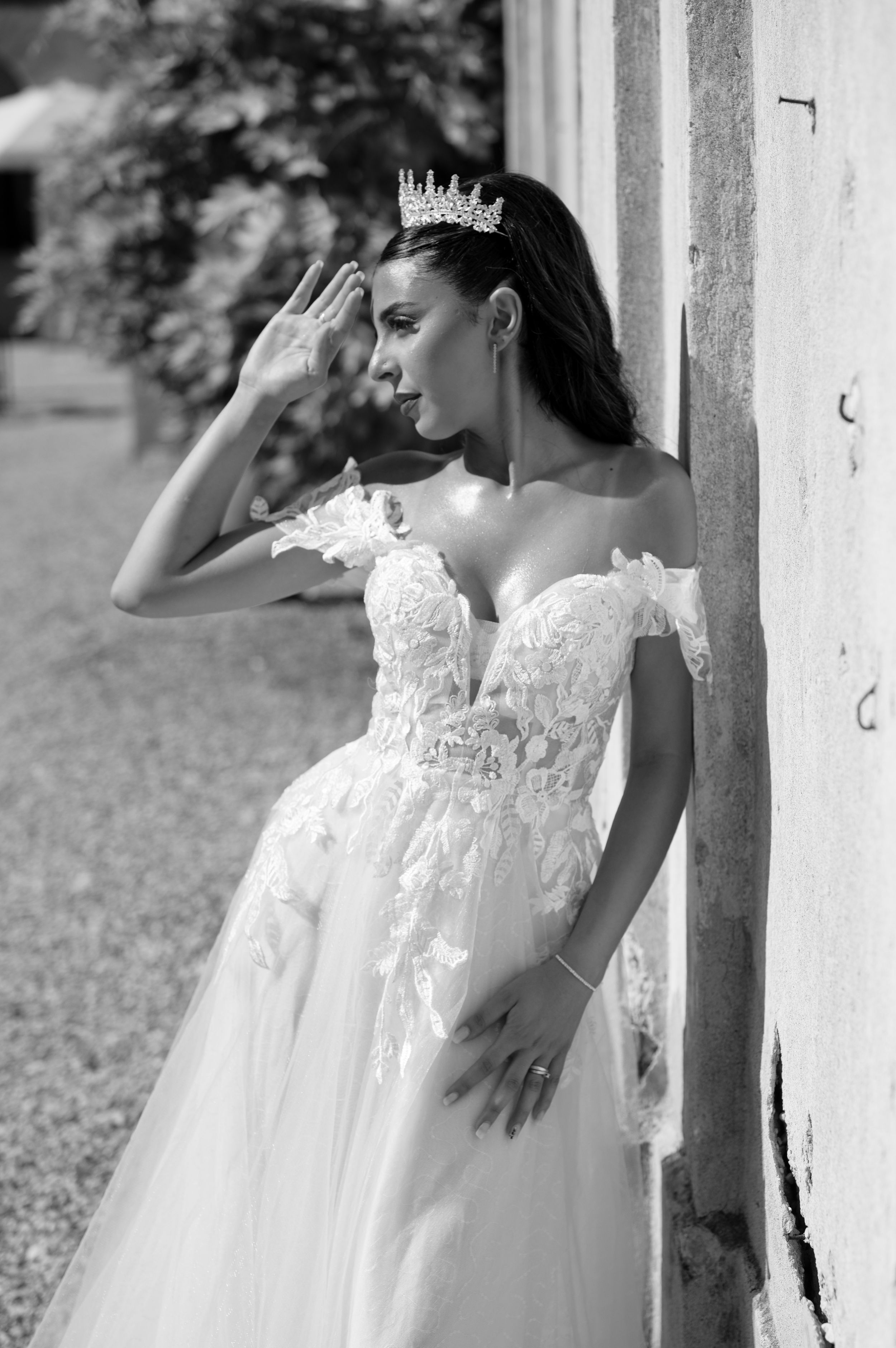 Bride in off-shoulder lace wedding gown and tiara poses gracefully against a wall in a black and white photo.