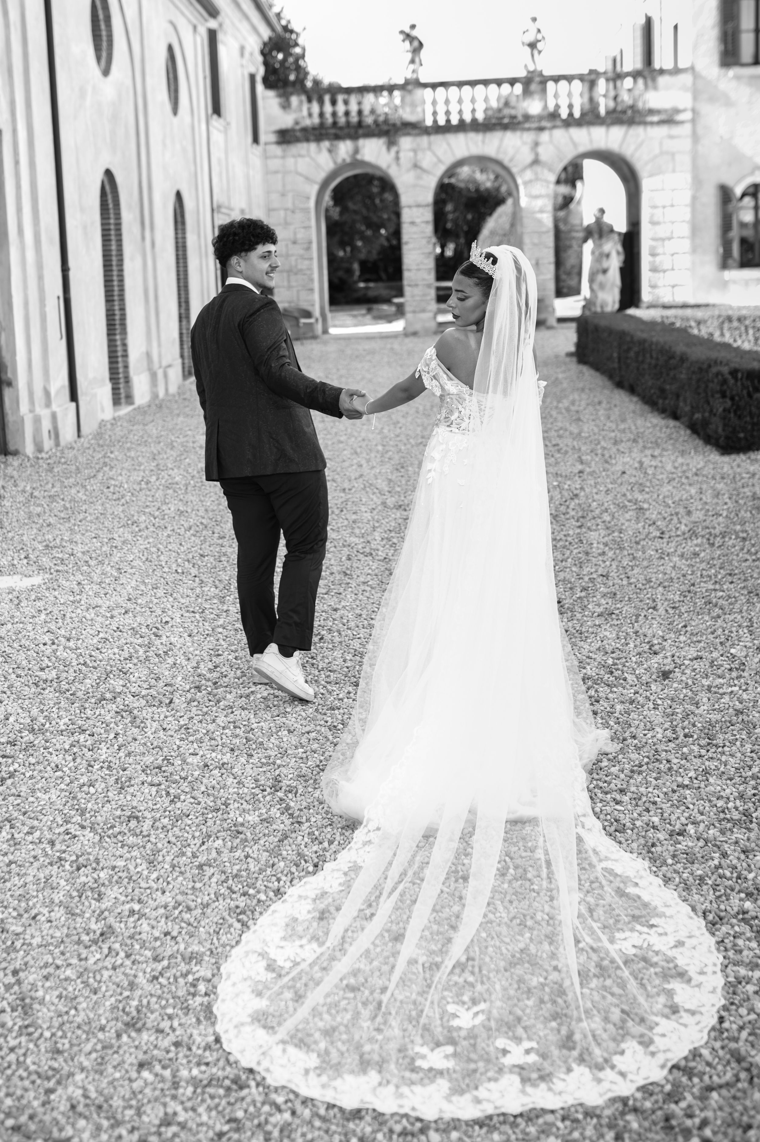 Black and white photo of bride and groom holding hands at elegant villa courtyard, long lace veil trailing.