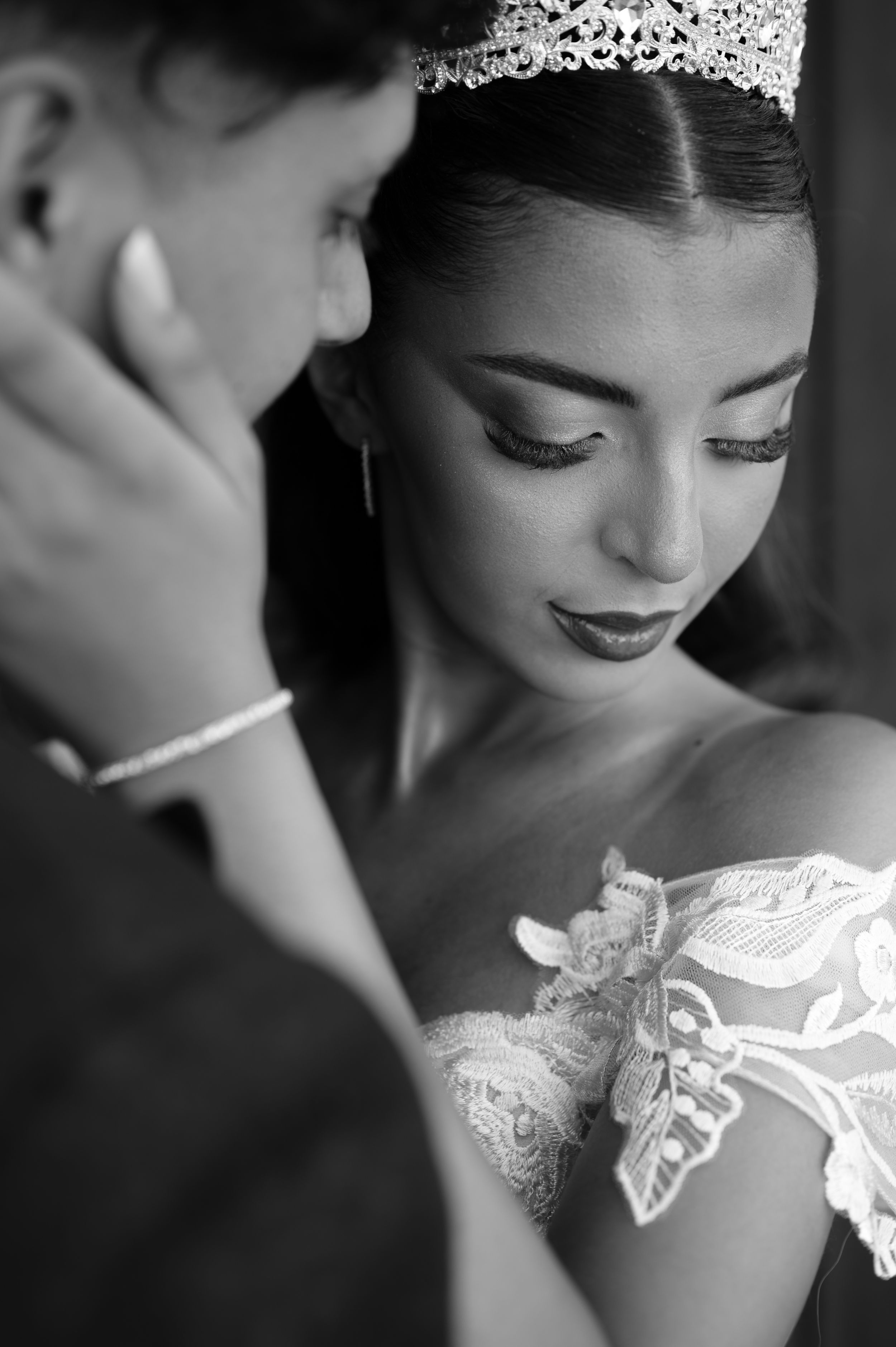 Black and white portrait of a bride in lace gown and tiara, groom tenderly touching her face.