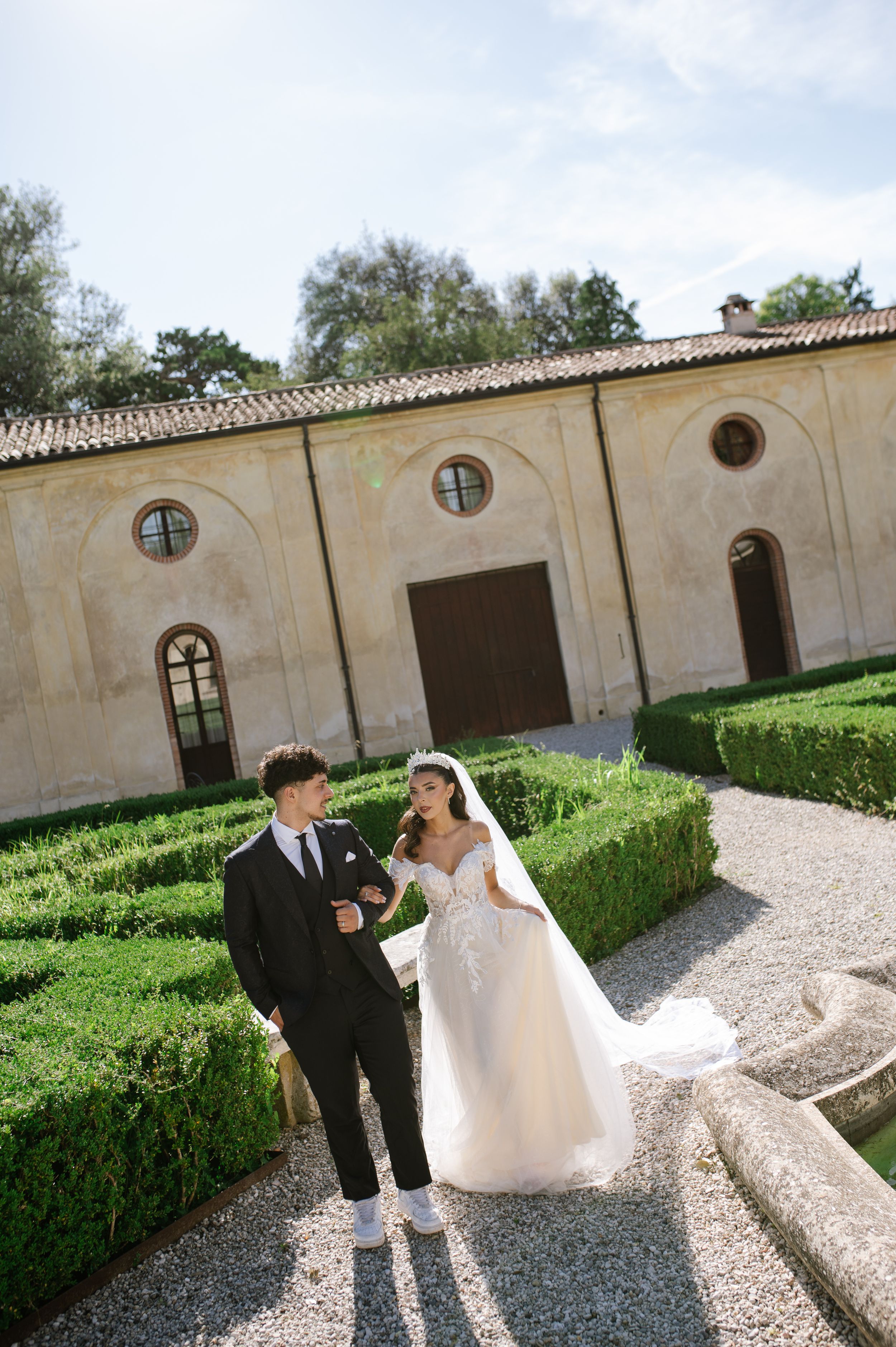 Bride and groom walk through elegant Italian villa garden with manicured hedges on their wedding day.