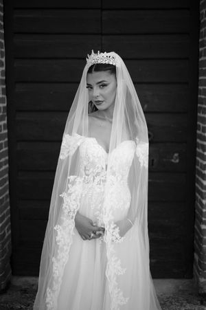 Black and white portrait of bride in lace wedding gown with crystal crown and cathedral veil before wooden doors.
