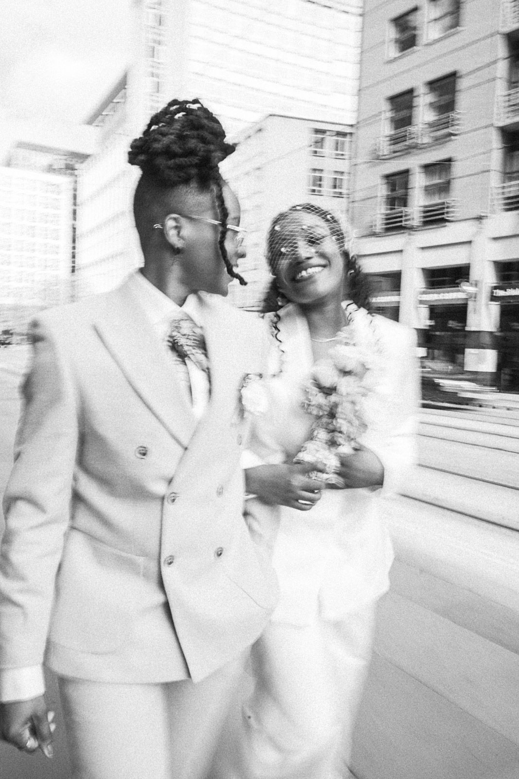 Black and white photo of two women in white suits smiling and walking together on a city street, one holding a bouquet.