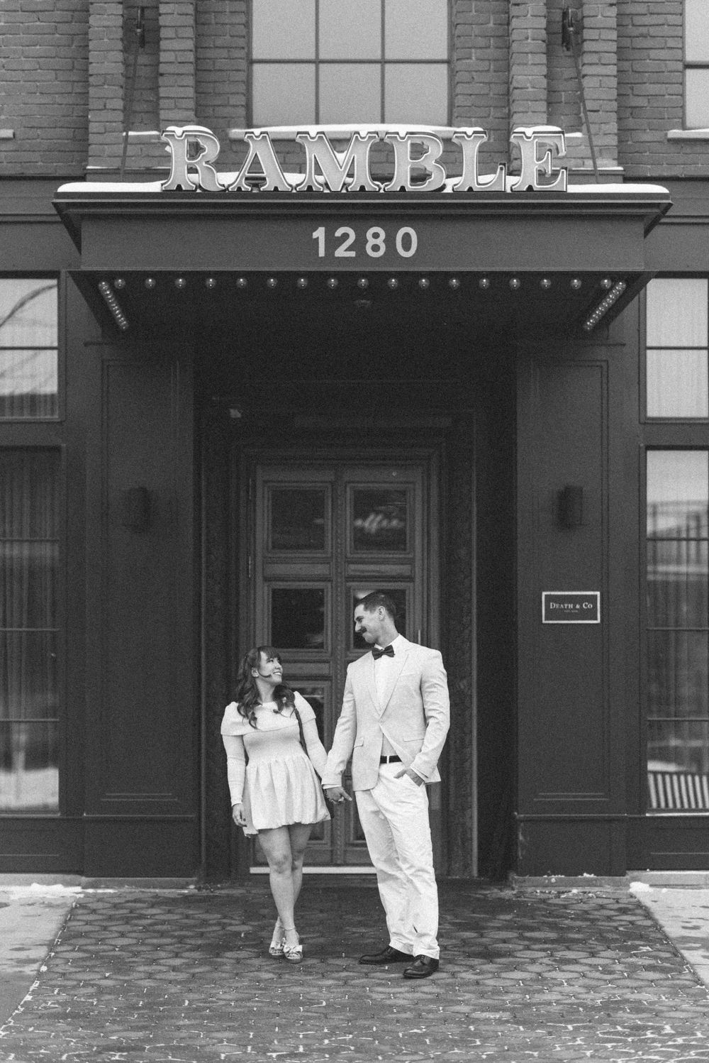 A couple stands beneath the Ramble sign outside a brick building entrance.