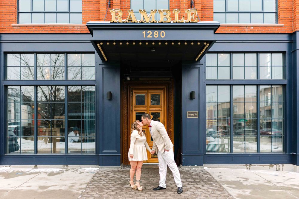 A couple poses in front of a brick building with large windows and an elegant black awning entrance.
