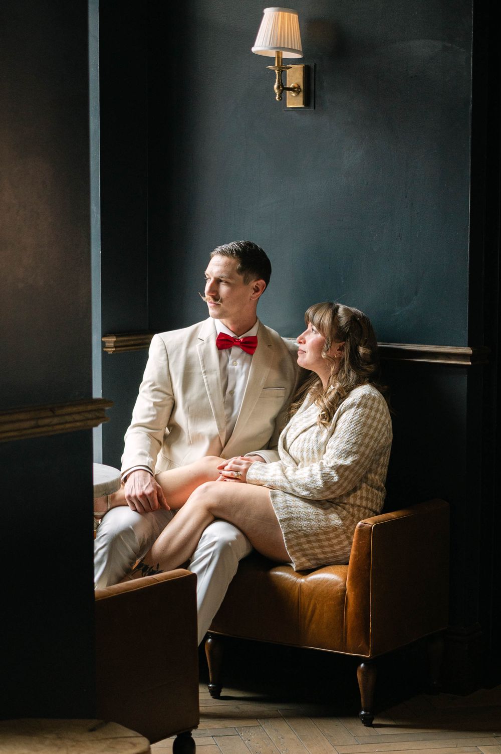 Couple in formal attire sharing intimate moment on leather couch in moody room with wall sconce.