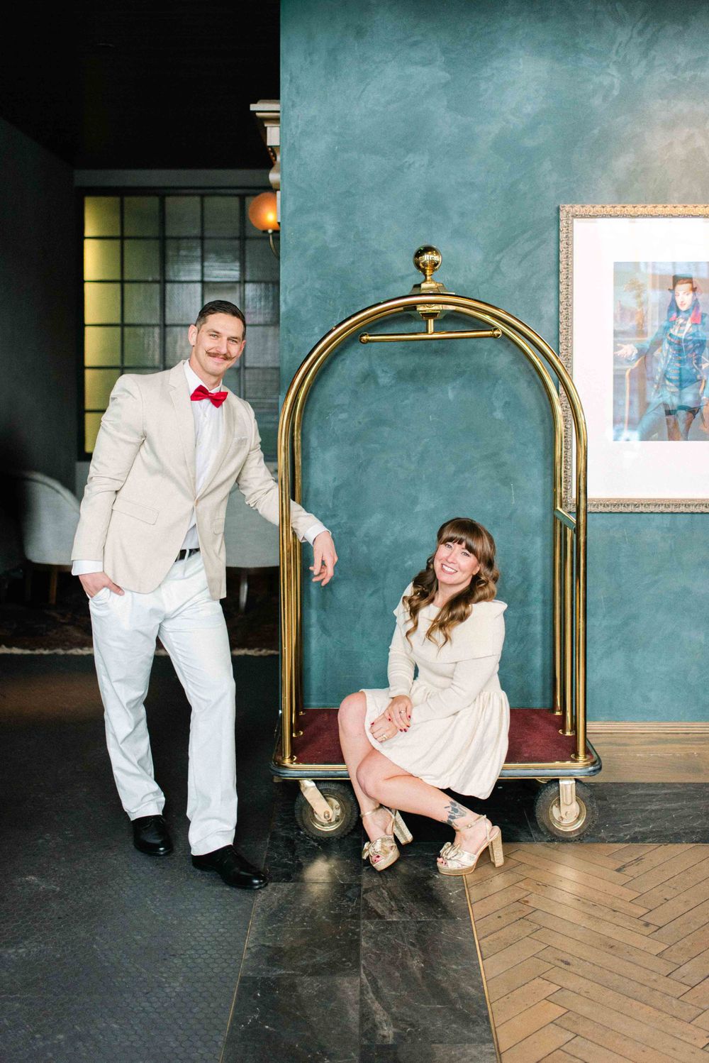 A couple poses playfully with a vintage hotel luggage cart against a teal wall.