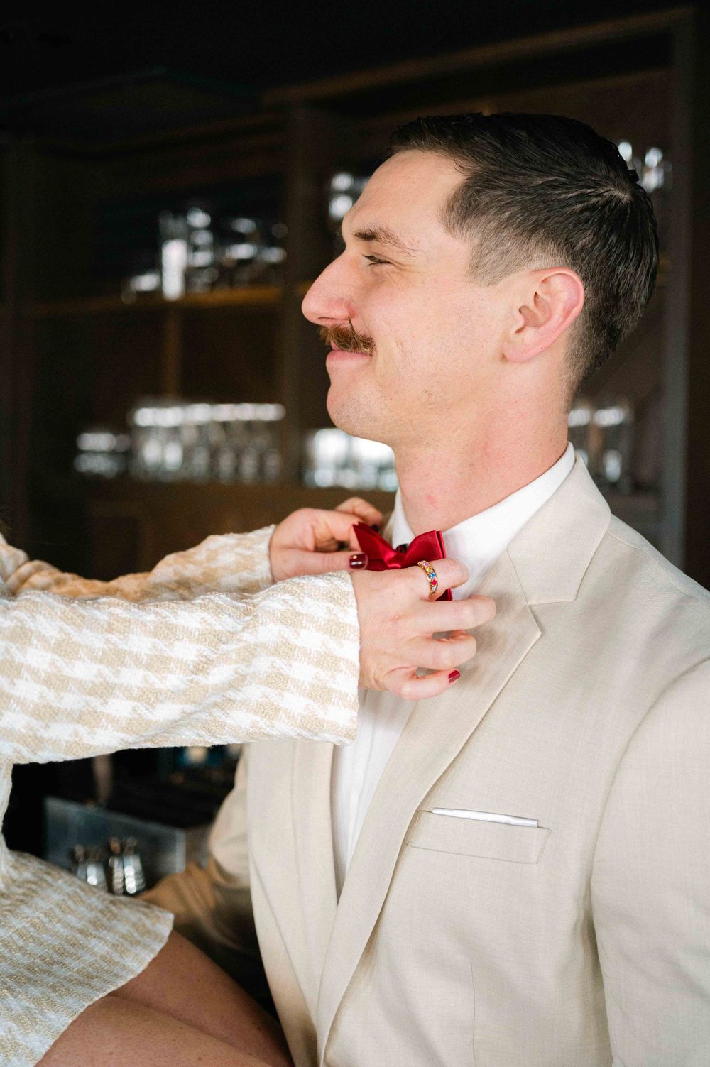A person in a white suit and red bowtie being helped with their tie adjustment.
