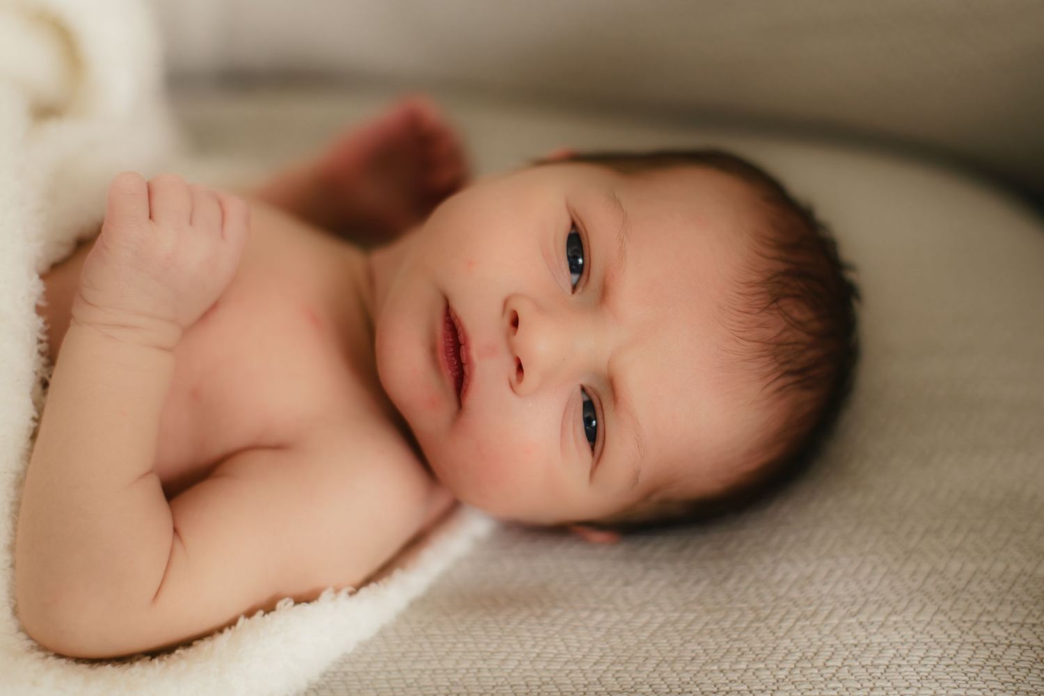 Newborn baby lying on soft beige fabric with alert expression and dark hair.