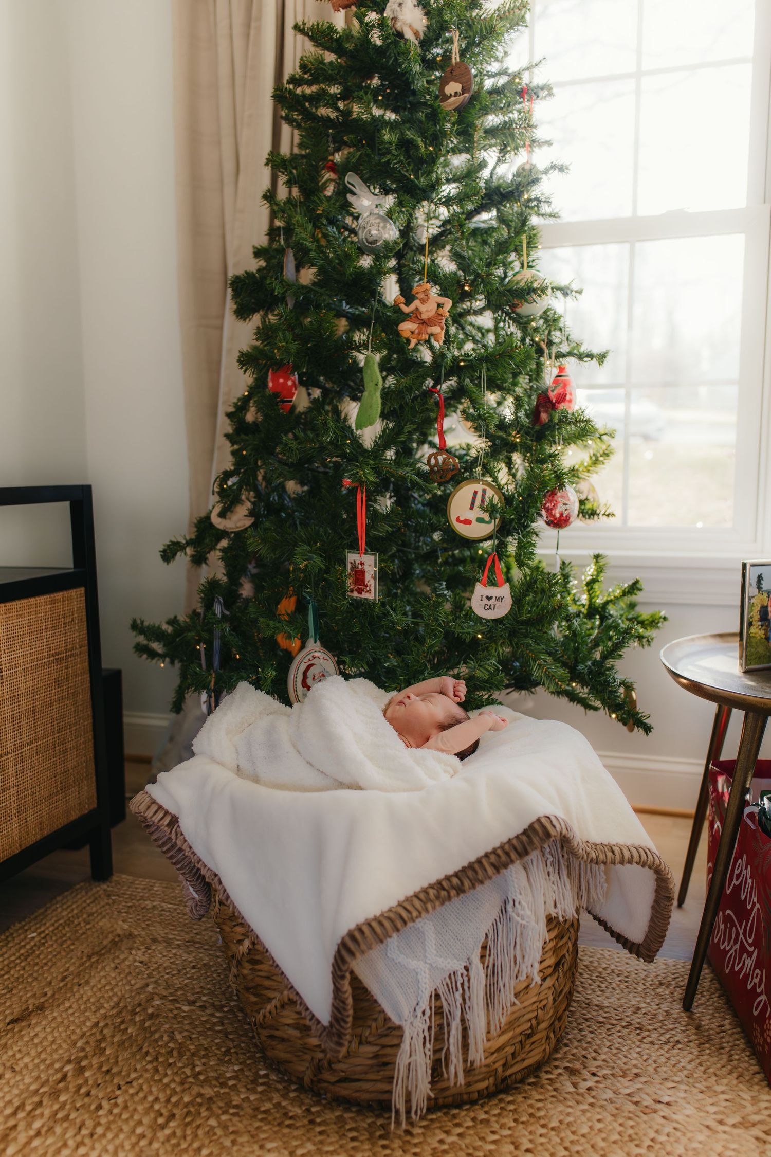 Newborn resting in white basket beneath decorated Christmas tree.