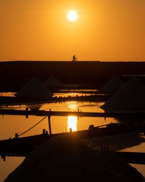 Silhouette of a cyclist on a hill against an orange sunset sky reflecting in salt ponds below.