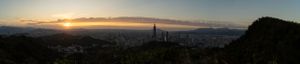 Panoramic cityscape view at sunset with mountains and urban buildings silhouetted against an orange sky.