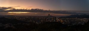 City skyline silhouetted against a dramatic sunset with dark clouds and mountain backdrop.