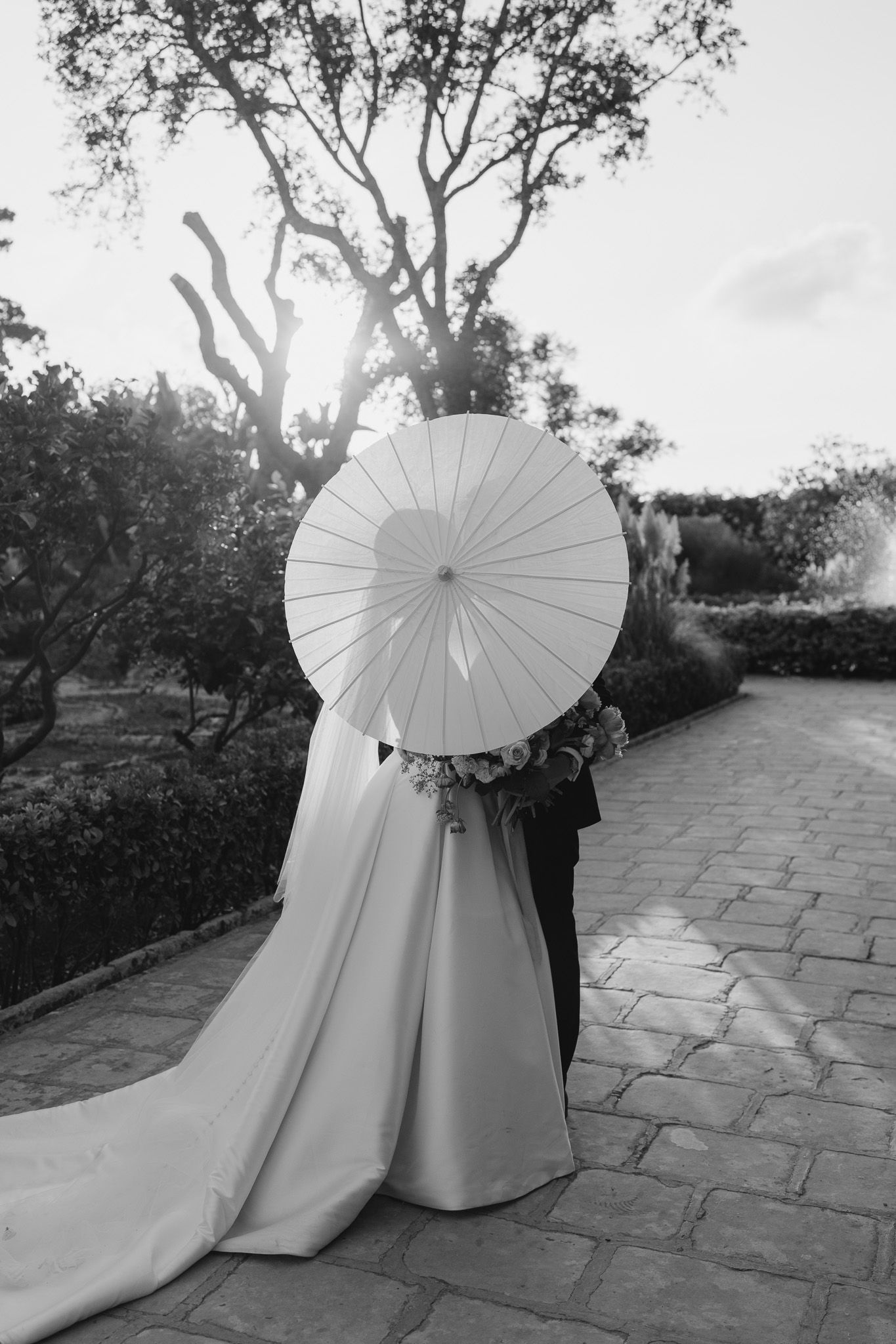 A wedding couple poses with a white parasol on a stone pathway.