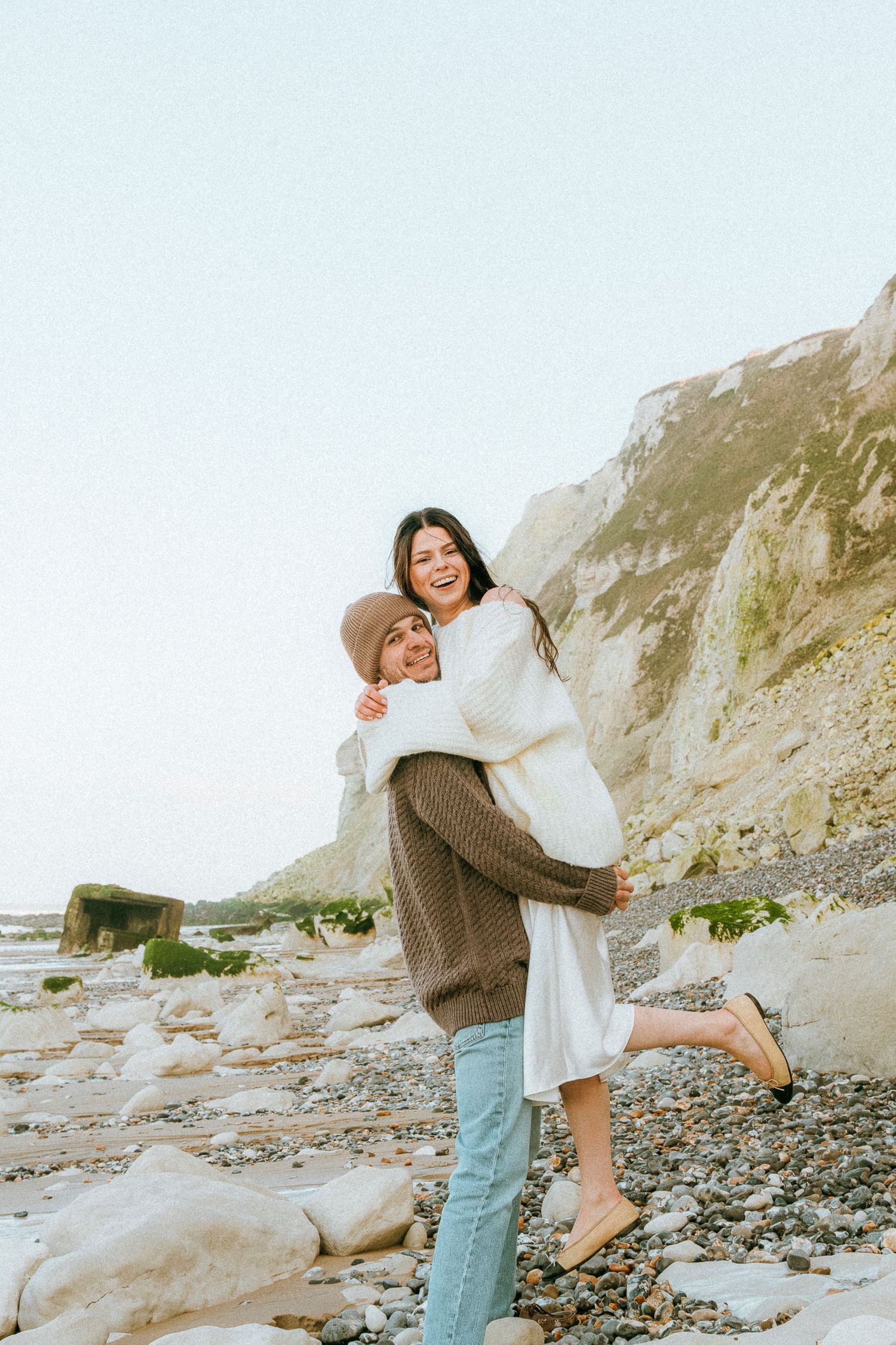 A couple embraces joyfully on a rocky beach with dramatic coastal cliffs in the background.