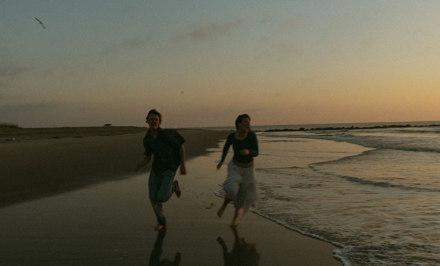 A couple holds hands while dancing on a beach at sunset with ocean waves and golden light in the background.