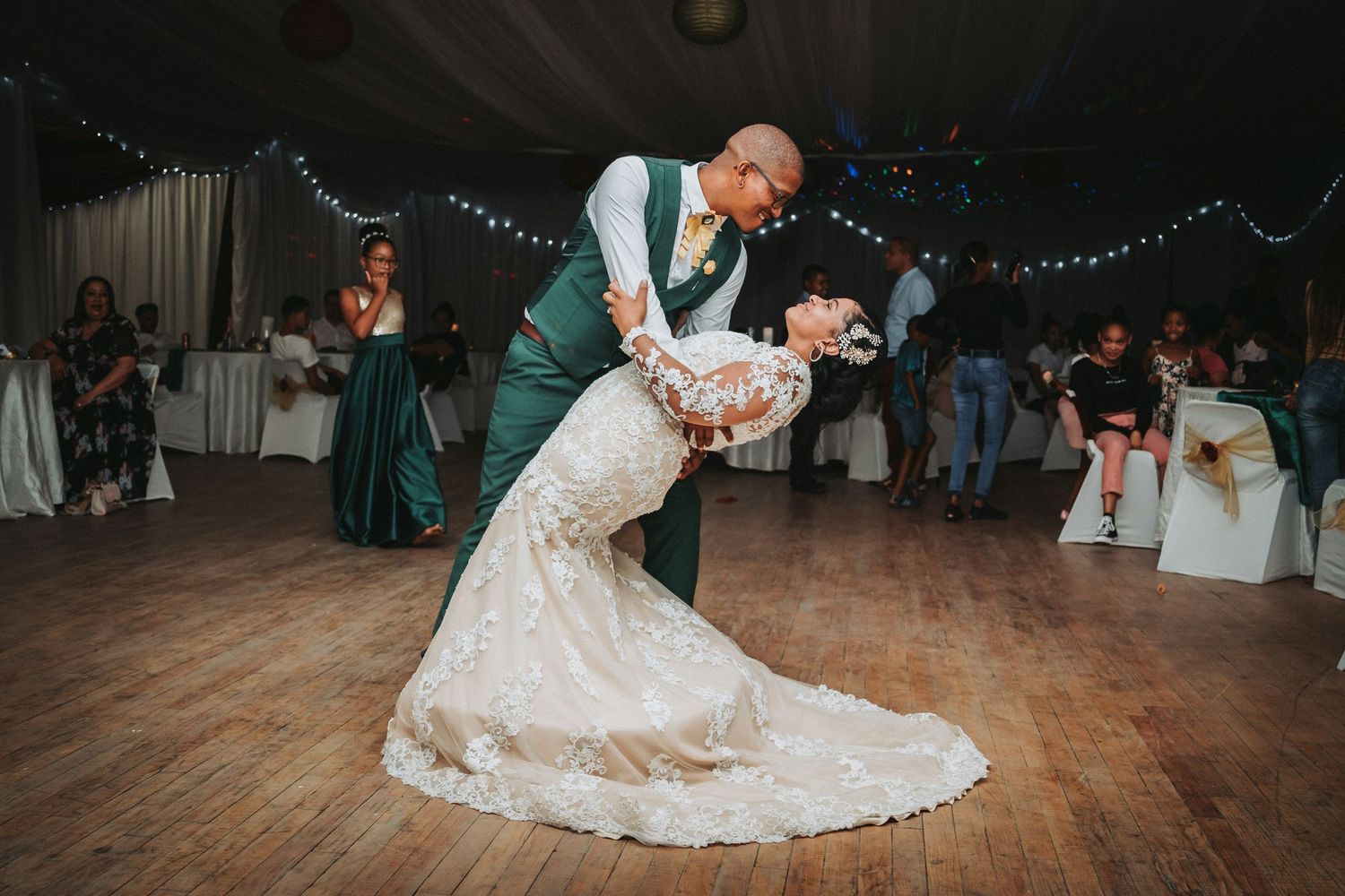 A couple performs an elegant dip on the wooden dance floor during their wedding reception, with guests watching in the background.