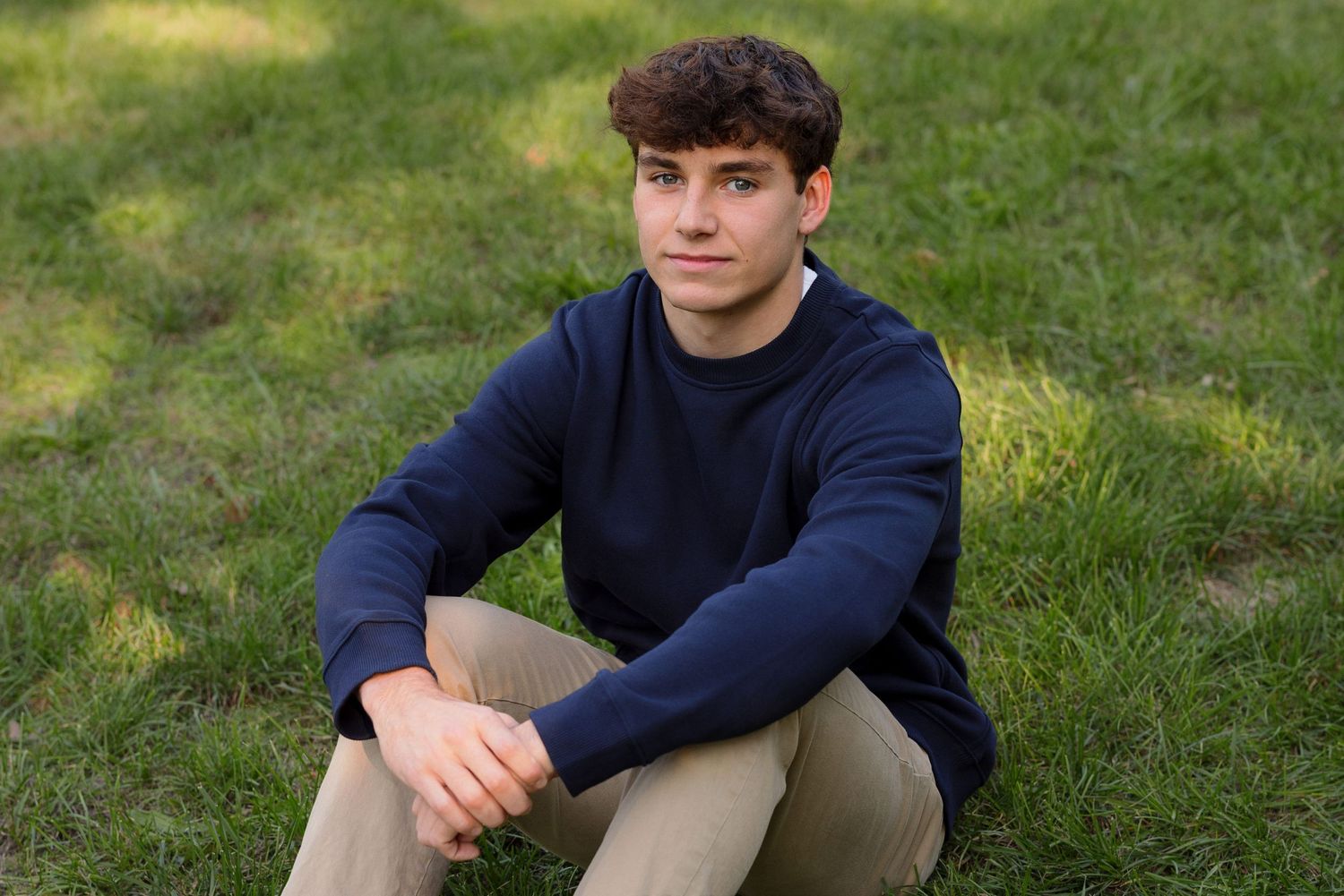 A person sits on grass wearing a navy blue sweater and khaki pants in a casual outdoor photo.