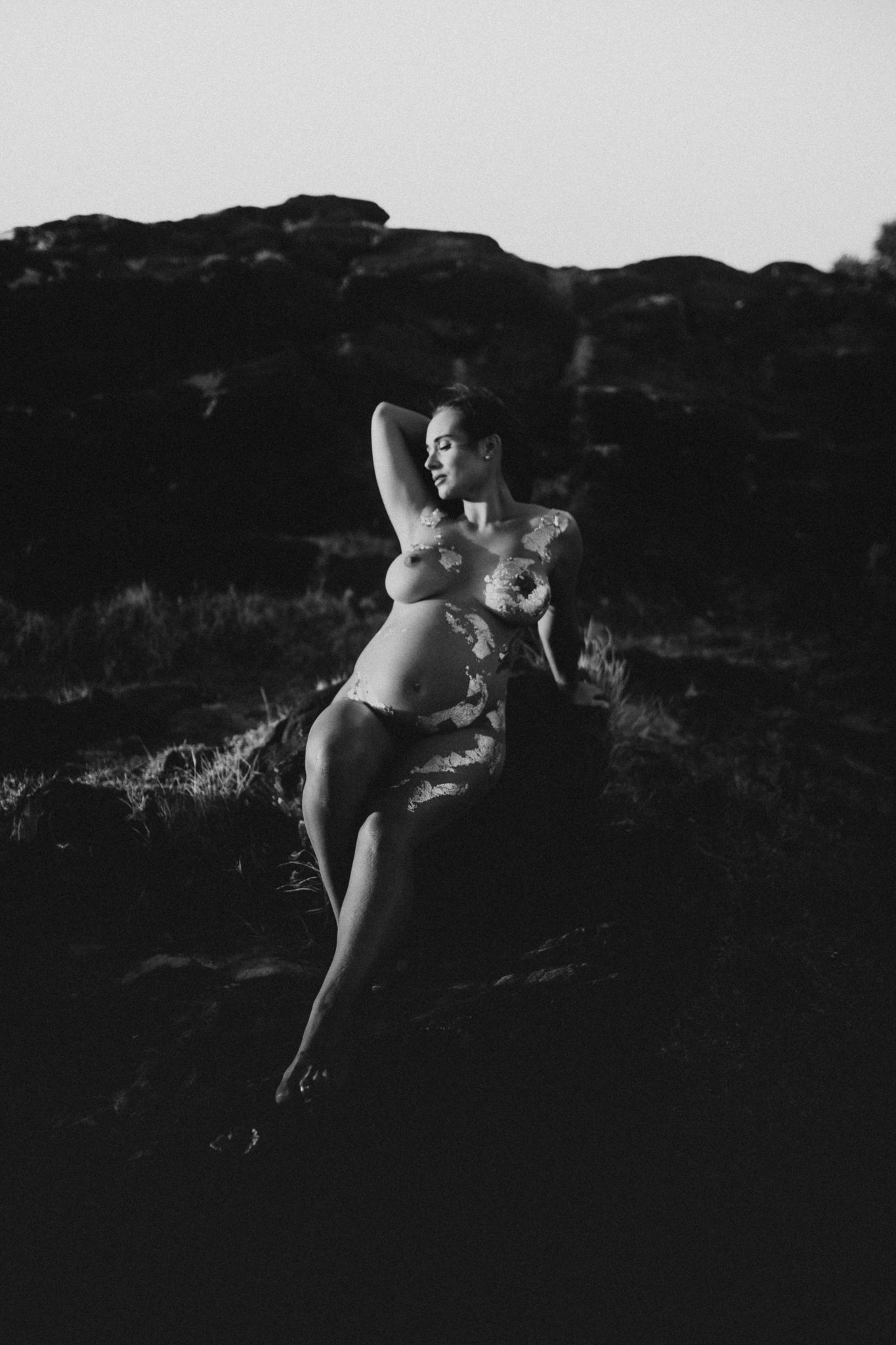 A dramatic black and white artistic nude photograph of a figure reclining against dark rocks in silhouette.
