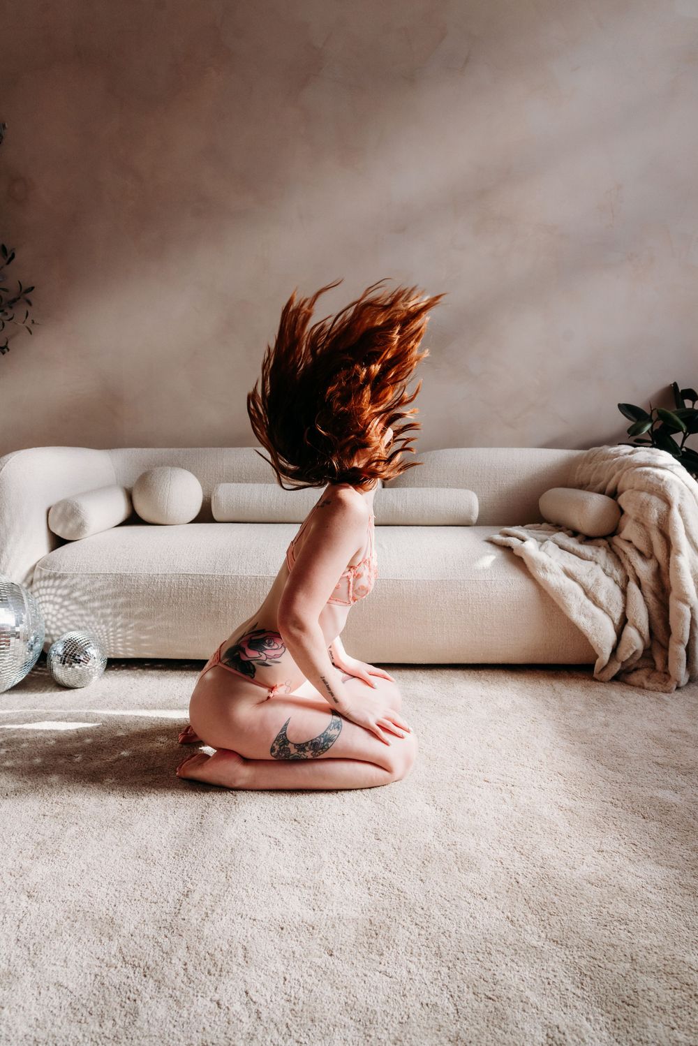 A person with red hair flipping their head back while kneeling on a beige carpet in front of a light colored couch.