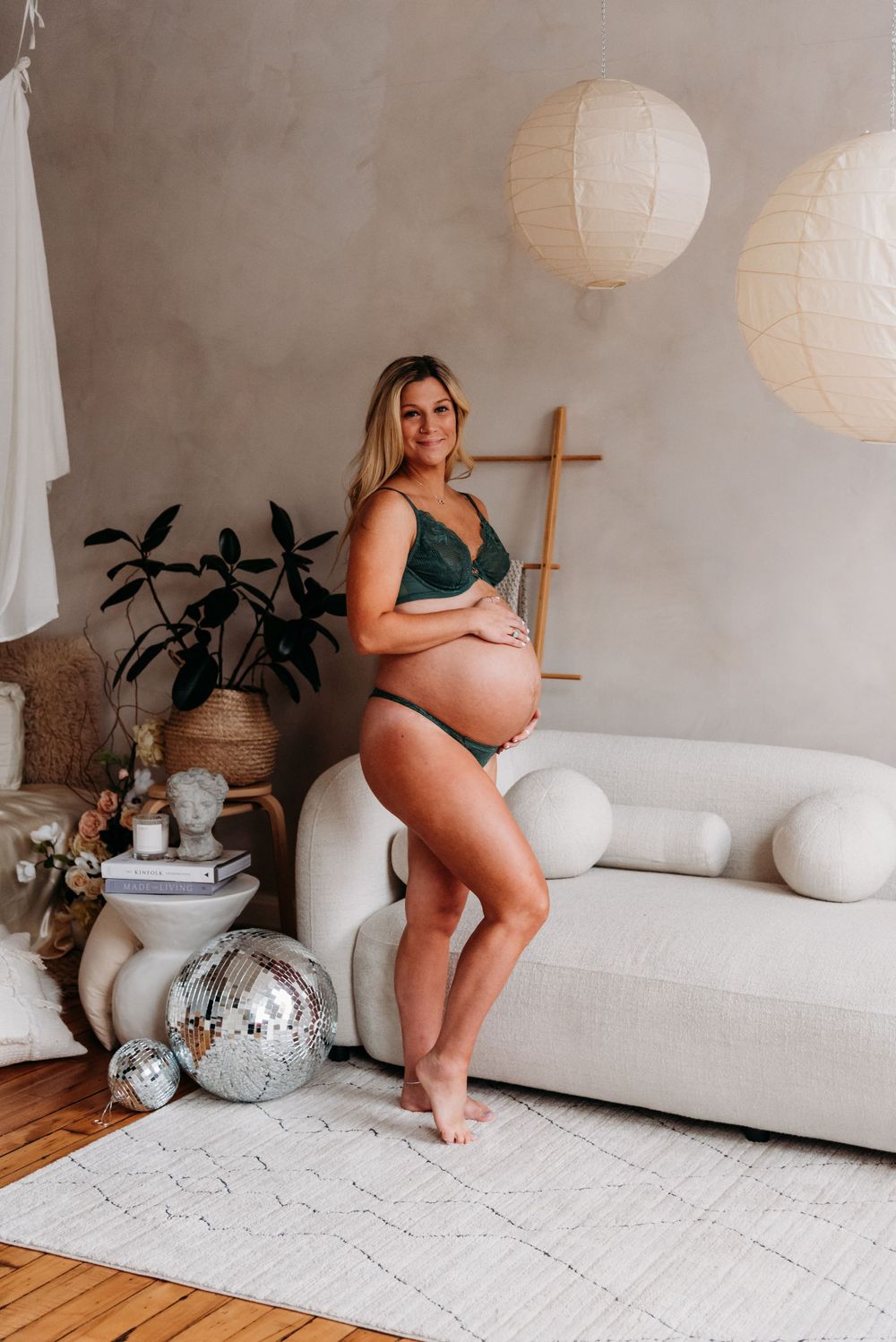 Maternity photo with paper lanterns, silver disco balls, and white couch in minimalist room decorated with potted plants.