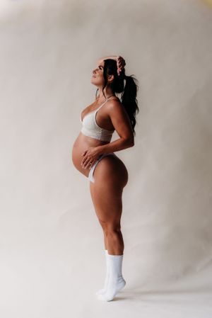 Artistic maternity photoshoot of expecting mother in white lingerie posing against neutral background.
