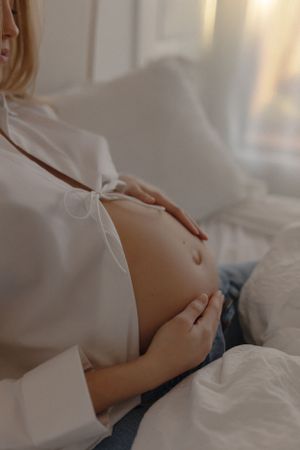Woman in white shirt caressing pregnant belly while relaxing on white bedding in soft natural light.