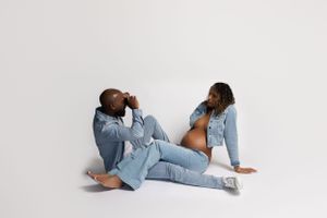 Two people wearing matching light denim outfits sitting and posing on white background in studio setting.