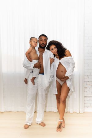 An elegant maternity photo featuring a family in white outfits against a light background.