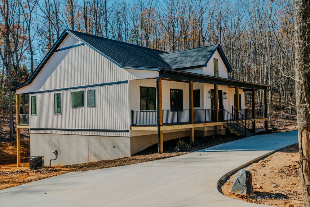 Modern farmhouse exterior with wraparound porch and metal roof in wooded setting.
