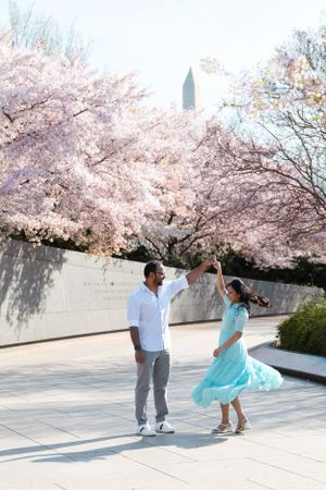 A couple dances playfully under blooming cherry blossom trees on a sunny spring day.
