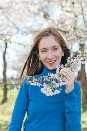 Person in bright blue sweater poses with cherry blossoms against blue sky.