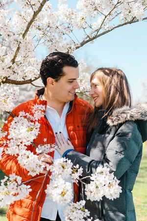 Couple in orange jacket and gray coat share tender moments among cherry blossoms.