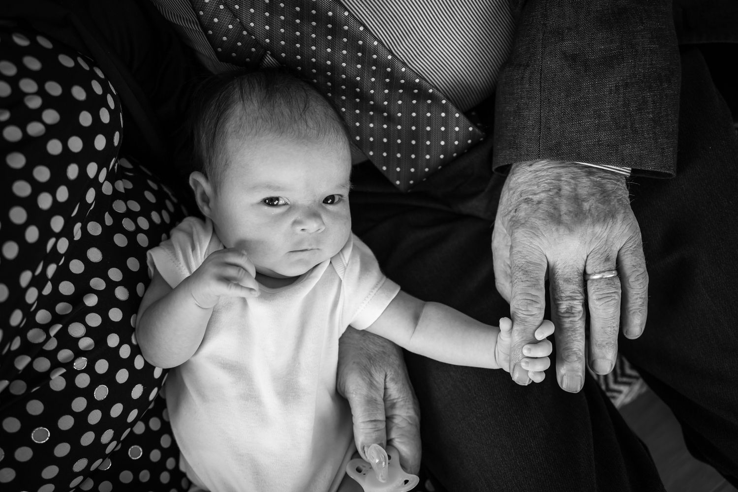 A black and white image shows a baby in a white outfit holding hands with someone wearing a polka dot pattern.