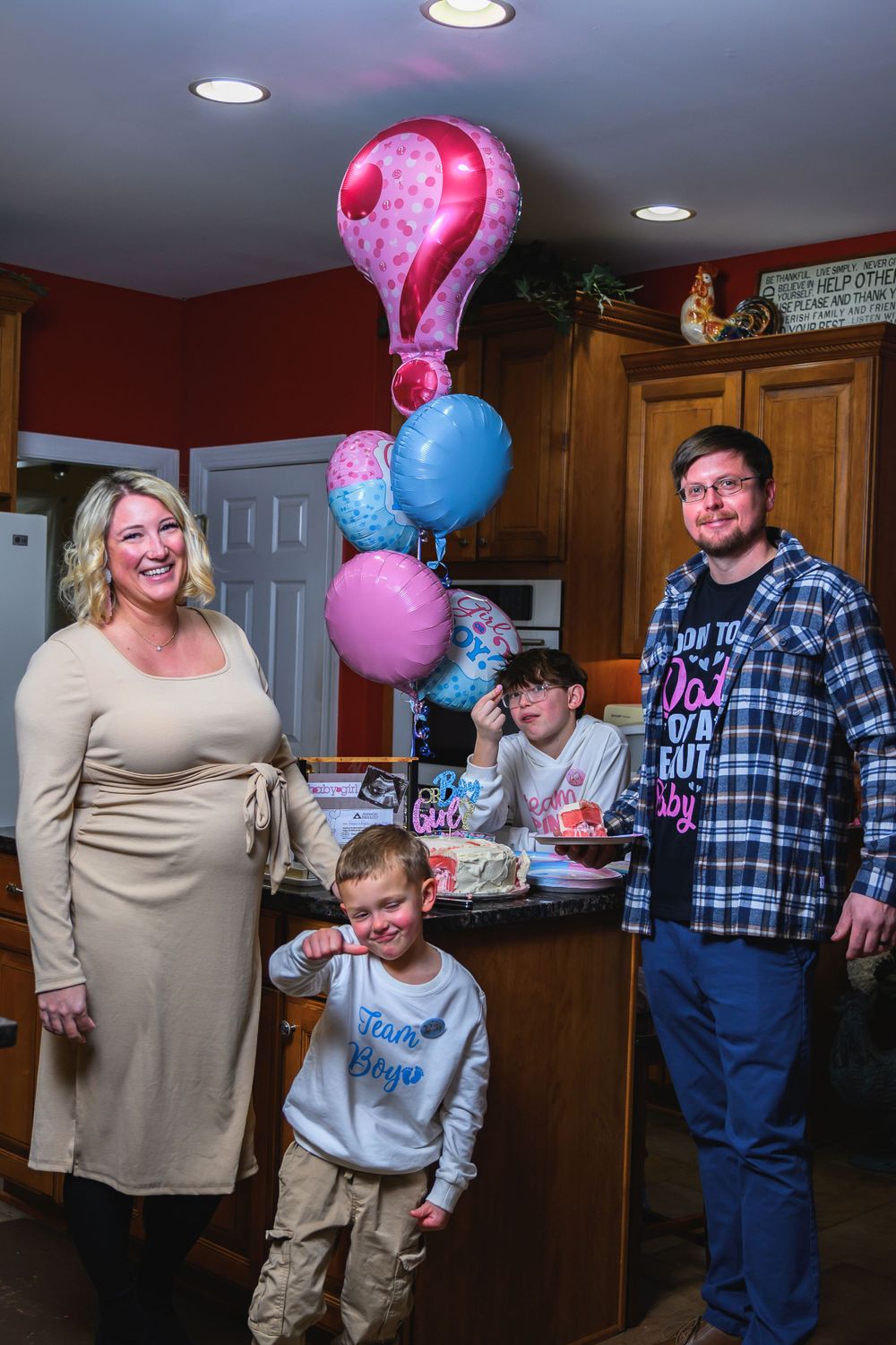 A group gathers in a kitchen for a celebration with pink and blue balloons featuring a large question mark balloon decoration.