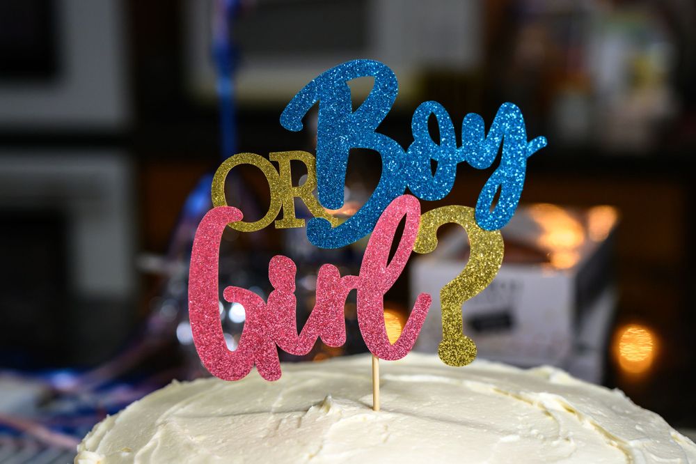 A glittery cake topper reading 'Boy or Girl' in pink and blue colors sits atop a white frosted cake.