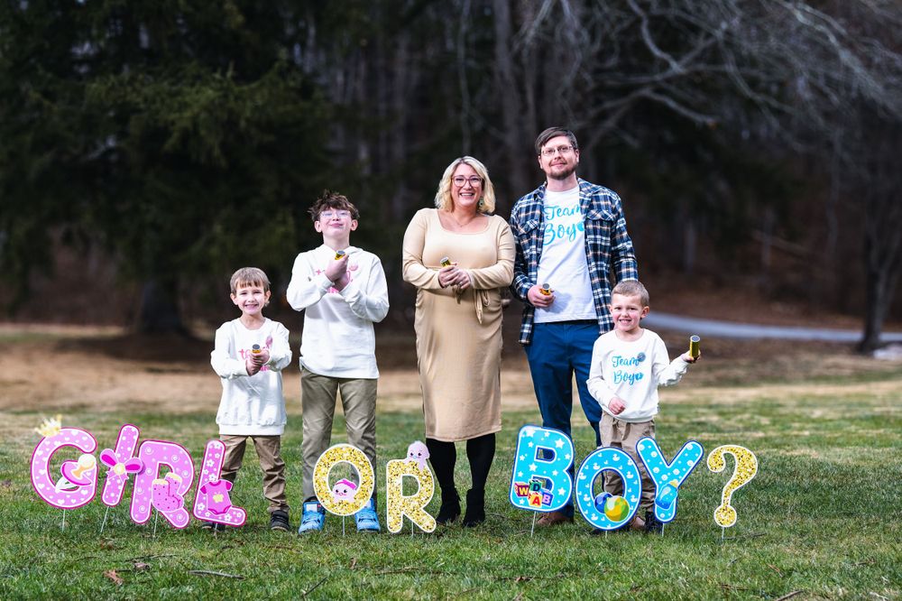 Family stands behind colorful 'GIRL OR BOY?' gender reveal lettering display set up outdoors on grass.